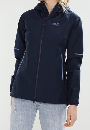 Frau trägt eine dunkelblaue Jack Wolfskin Kapuzenjacke mit Frontreißverschluss und hellblaue zerrissene Jeans vor weißem Hintergrund.