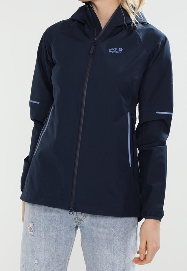 Femme portant une veste à capuche Jack Wolfskin bleu marine avec fermeture éclair à l'avant et un jean clair déchiré sur un fond blanc.