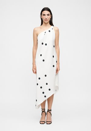 Kobieta stojąca w białej sukience z jednym ramieniem w czarne grochy, nosząca czarne sandały na wysokim obcasie na jednolitym tle.