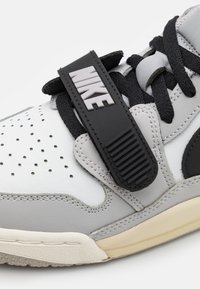 Šedé a bílé tenisky s perforovaným designem, černými akcenty a výrazným černým pásem s logem "NIKE" na boku.