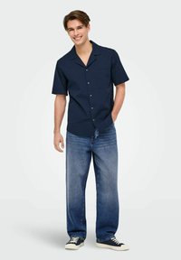 Ung mann står, smilende, iført en marineblå kortermet skjorte med knapper, blå jeans og marineblå sneakers med hvite såler.