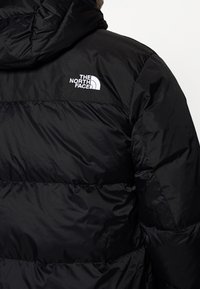 Gros plan d'une personne portant une doudoune noire avec une capuche et un logo blanc "The North Face" sur le haut du dos.