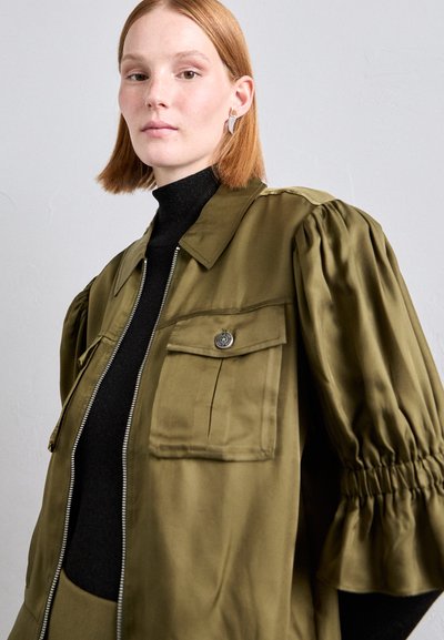 Olívovozelená oversized bunda so zipsom vpredu, zapínacou náprsníkovou vreckou a nadúvacími rukávmi, vrstvená nad čiernym topom s vysokým golierom.
