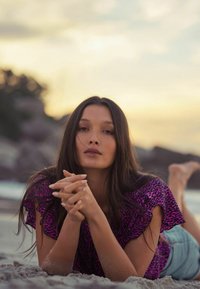 Modèle portant un haut en imprimé léopard violet vibrant avec des manches volantées, allongée sur une plage de sable, avec une peau bronzée et de longs cheveux noirs.
