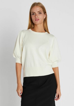 Mladá žena s dlhými blond vlasmi, oblečená v bielom pulóvri s nafúknutými rukávmi a čiernej sukni, pózujúca pred jednoduchým svetlým pozadím.