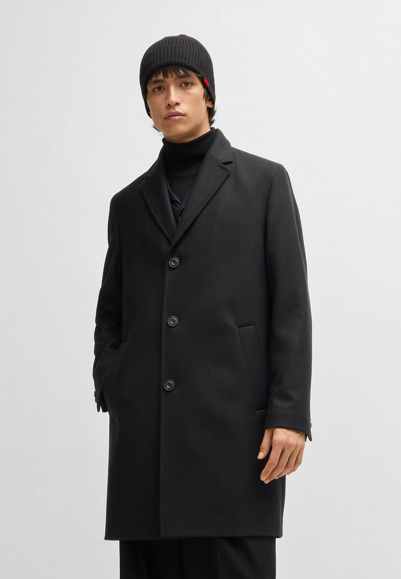 Black wool overcoat with a notched lapel, four buttons, and side pockets. Model wears a ribbed black beanie and a black turtleneck underneath.
