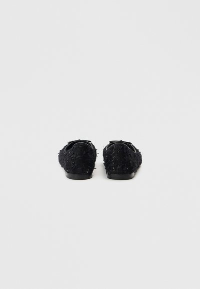 Μαύρα παπούτσια τύπου slip-on με υφή ύφασμα, διακοσμημένα με λεπτομέρειες κρόσσια, στρογγυλεμένη μύτη, χαμηλό τακούνι και μινιμαλιστική μαύρη σόλα.