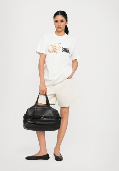 Tricou grafic alb cu un design de ceașcă de ceai și cuvântul "TRICOU", asociat cu pantaloni scurți deschisi, o geantă de piele neagră și balerini negri.