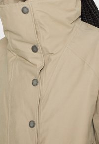 Veste beige à col montant avec boutons-pression métalliques et coutures visibles sur les épaules et les panneaux avant.