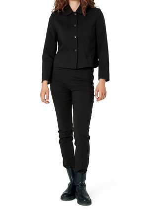 Kobieta w czarnej marynarce na guziki, dopasowanych czarnych spodniach i czarnych botkach do kostek stoi na tle białym.