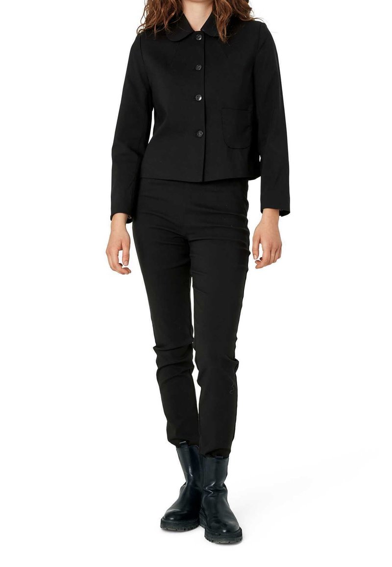 Kobieta w czarnej marynarce na guziki, dopasowanych czarnych spodniach i czarnych botkach do kostek stoi na tle białym.