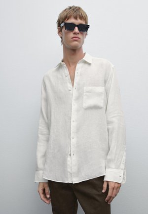 Camicia bianca in lino a maniche lunghe con taschino sul petto, apertura con bottoni e orlo leggermente curvo. Indossata con occhiali da sole neri a forma rettangolare.