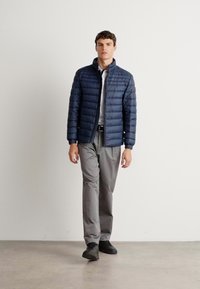 Man walking indoors wearing a navy quilted jacket, white shirt, gray trousers, black belt, and black shoes against a plain white wall.
