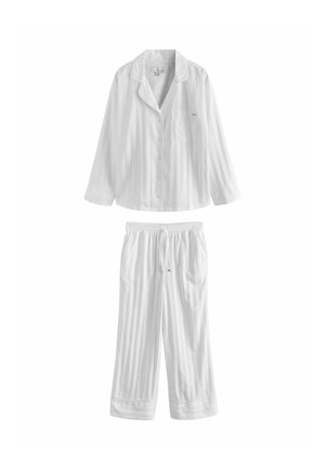 Hosszú hajú nő, aki laza fehér, gombos inget és bőszárú nadrágot visel, mezítláb áll egy texturált, halvány rózsaszín fal előtt.