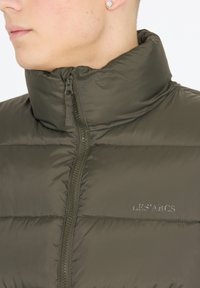Dunkelgrüne Pufferjacke mit hohem Kragen, glatter Textur, Reißverschluss vorne und geprägtem Logo "LES'ARCS" auf der Brust.