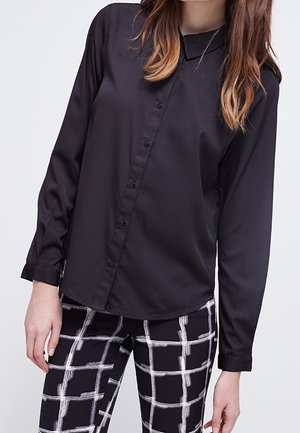 Mujer con una camisa negra de manga larga abotonada y pantalones negros con un patrón de cuadrícula blanca, de pie contra un fondo claro y liso.