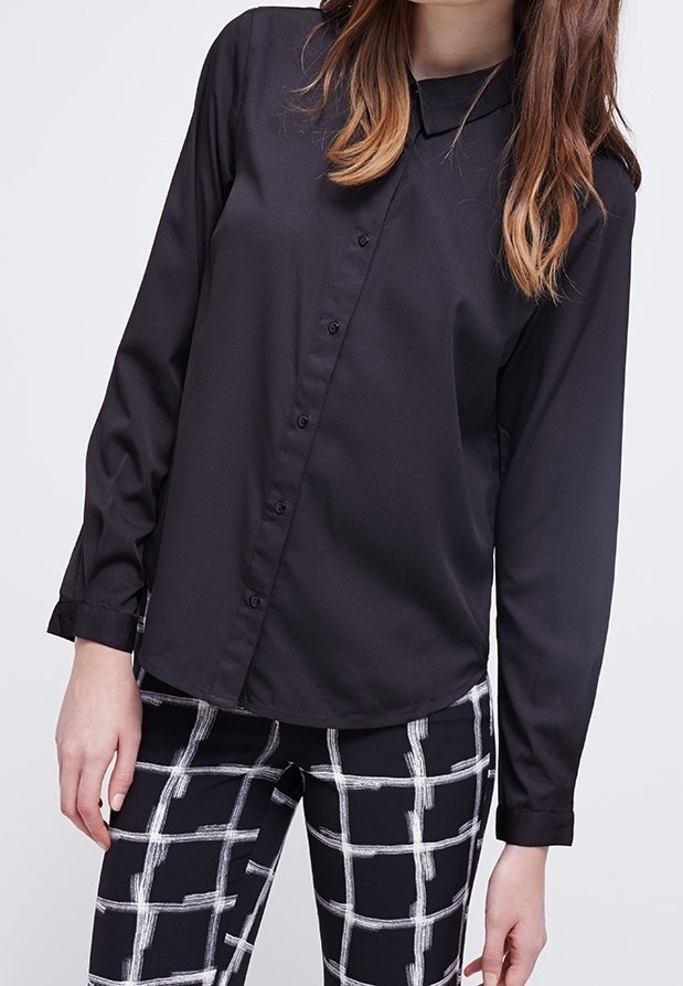 Femme portant une chemise noire à manches longues et pantalons noirs avec un motif à quadrillage blanc, debout devant un fond clair uni.