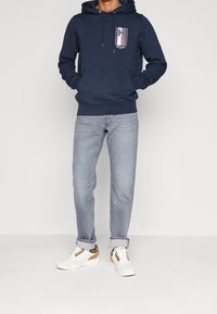 Sudadera azul marino con un bolsillo frontal, cordones blancos y un gráfico en el pecho. Combinada con jeans azul claro y zapatillas blancas con acentos marrones.