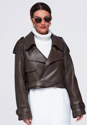 Femme portant une veste en cuir courte marron foncé sur un pull à col roulé blanc et des lunettes de soleil rondes sur un fond uni.