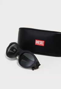 Lunettes de soleil noires avec un design arrondi, finition matte et verres foncés. Comprend un étui noir avec un logo "DIESEL" rouge.