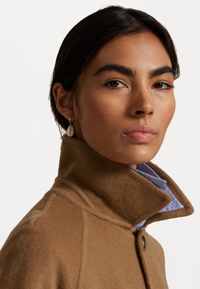 Brown wool coat with a high collar, featuring smooth texture and a button detail, worn over a light blue shirt. Gold earring accent.