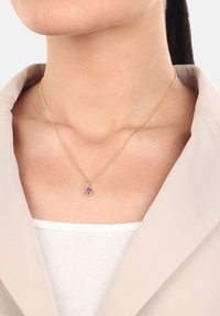 Collana in oro con pendente a forma ovale di pietra viola, indossata su un collo nudo. Lo sfondo è neutro con un blazer di colore chiaro.