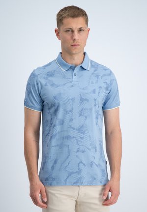 Man die een lichtblauw polo met patroon en beige broek draagt, staat voor een lichte effen achtergrond, recht vooruit kijkend.