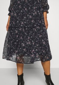 Robe midi noire transparente avec imprimé floral rose et blanc, manches longues et ourlet à volants, portée avec des bottines noires.