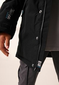 Giacca nera in tessuto liscio, con zip laterale, bottoni a pressione e coordinate stampate sulla manica. Pantaloni grigi visibili sotto.
