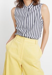 Randig ärmlös blus i marinblått och vitt, med knäppning framtill och ficka, kombinerad med högmidjade, ribbade gula byxor.