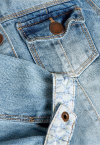 Tessuto in denim blu chiaro con cuciture marroni a contrasto. Presenta due tasche con chiusure a bottone metallico e un dettaglio in tessuto con motivo floreale.