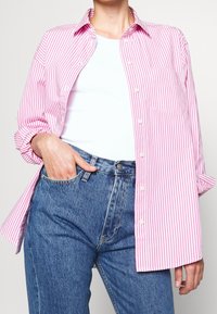 En rosa och vitrandig skjorta med knapp, med krage, bärs över en vit topp och kombineras med blå denimjeans; har en bröstficka.