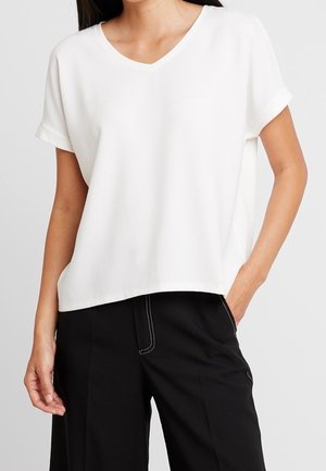 Kobieta ubrana w luźną białą bluzkę z krótkim rękawem, zestawioną z czarnymi szerokimi spodniami, jedną ręką w kieszeni, na jednolitym tle.