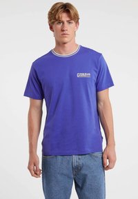 Młody mężczyzna ubrany w niebieską koszulkę z krótkim rękawem z logo "GUESS JEANS" oraz jasnoniebieskie dżinsy, stojący na tle białego tła.
