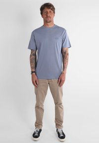 T-shirt en coton bleu clair à manches courtes, avec un logo sur la poitrine, accompagné d'un pantalon beige et de baskets noires, dotées de lacets blancs.
