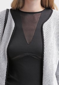 Femme portant un haut noir avec un panneau en V transparent et une veste blanche texturée avec un motif nid d'abeilles sur les manches et l'avant.