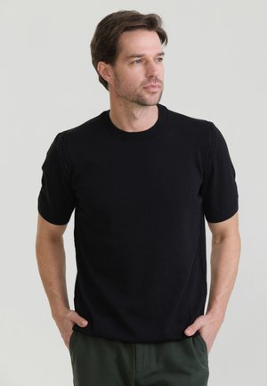 Mand med kort brunt hår iført en sort kortærmet sweater og mørkegrønne bukser, står med hænderne i lommerne og kigger til siden.