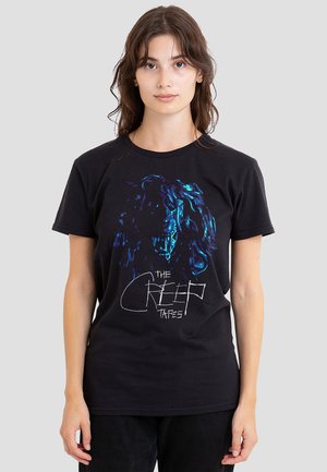 Young woman stands facing forward wearing black T-shirt with blue wolf face graphic and handwritten text "The Creep Tapes" on front.
