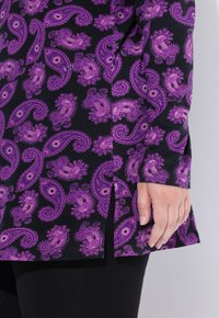 Zwarte kleding met paars paisleypatroon, gemaakt van een textuurstof. Zijdelingse splitdetails zichtbaar, tegen een neutrale achtergrond.