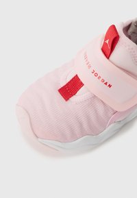 Scarpa sportiva rosa con upper in tessuto morbido, dotata di una cinghia rossa e logo. Suola bianca con dettagli testurizzati verso la punta.
