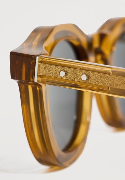 Gros plan sur des lunettes de soleil couleur ambre avec des montures épaisses translucides et des branches métalliques dorées avec deux petits rivets.