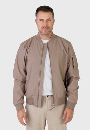 Homme portant une veste aviateur marron clair par-dessus une chemise blanche, debout devant un fond clair uni.
