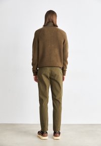Pull en maille marron avec col montant, pantalon vert et baskets marron. Accent sur les textures et palette de couleurs sourdes.