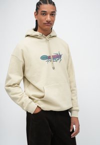 Beige hoodie met een voorzak, voorzien van een multikleurig grafisch ontwerp waarin de woorden "Volcano Stone Conservation" staan.