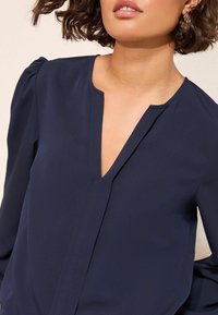 Jūras zils blūze ar V veida kakla izgriezumu un garām piedurknēm, kam ir smalkas plecu krokas. Gluda auduma struktūra ar tīru dizainu un minimālu detaļu klātbūtni.