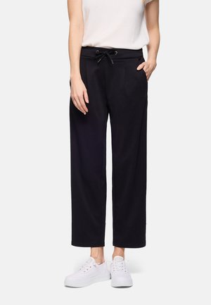 Mujer con pantalones negros de pierna ancha y cintura con cordón, y zapatillas blancas, de pie con una mano en el bolsillo.