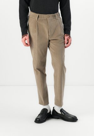 Pantaloni din corduroy de culoare maro deschis cu o croială largă, bandă elastică la talie și buzunare laterale, asortați cu mocasini negri și șosete albe.