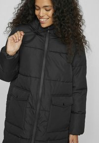 Melnā polsterētā jaka ar rāvējslēdzēju, kapuci ar šnauri un divām frontālajām kabatām. Iespējas ar quilted dizainu un brīvu piegriezumu. Mīksta tekstūra.