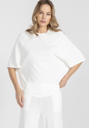 Kobieta z falującymi blond włosami, ubrana w luźną, białą bluzkę z krótkim rękawem i pasujące białe spodnie na jednolitym szarym tle.