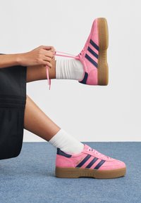 Person som knyter rosa Adidas-sneakers med mörkblå ränder och bruna sulor, iförd vita strumpor och en svart kjol, mot en enfärgad vit bakgrund.